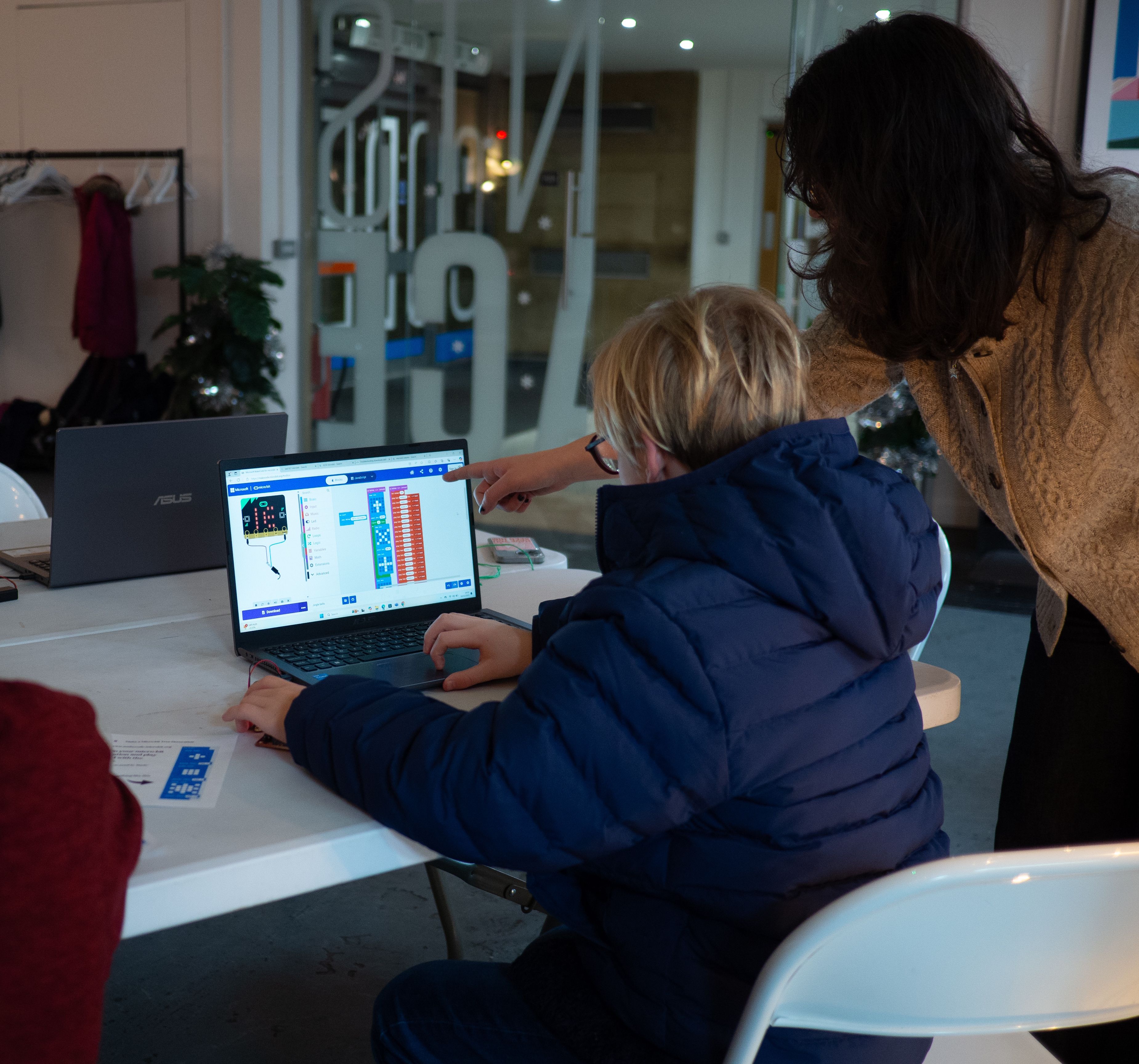 An attendee is programming a micro:bit on a laptop and a mentor is standing next to him, pointing at something on the screen