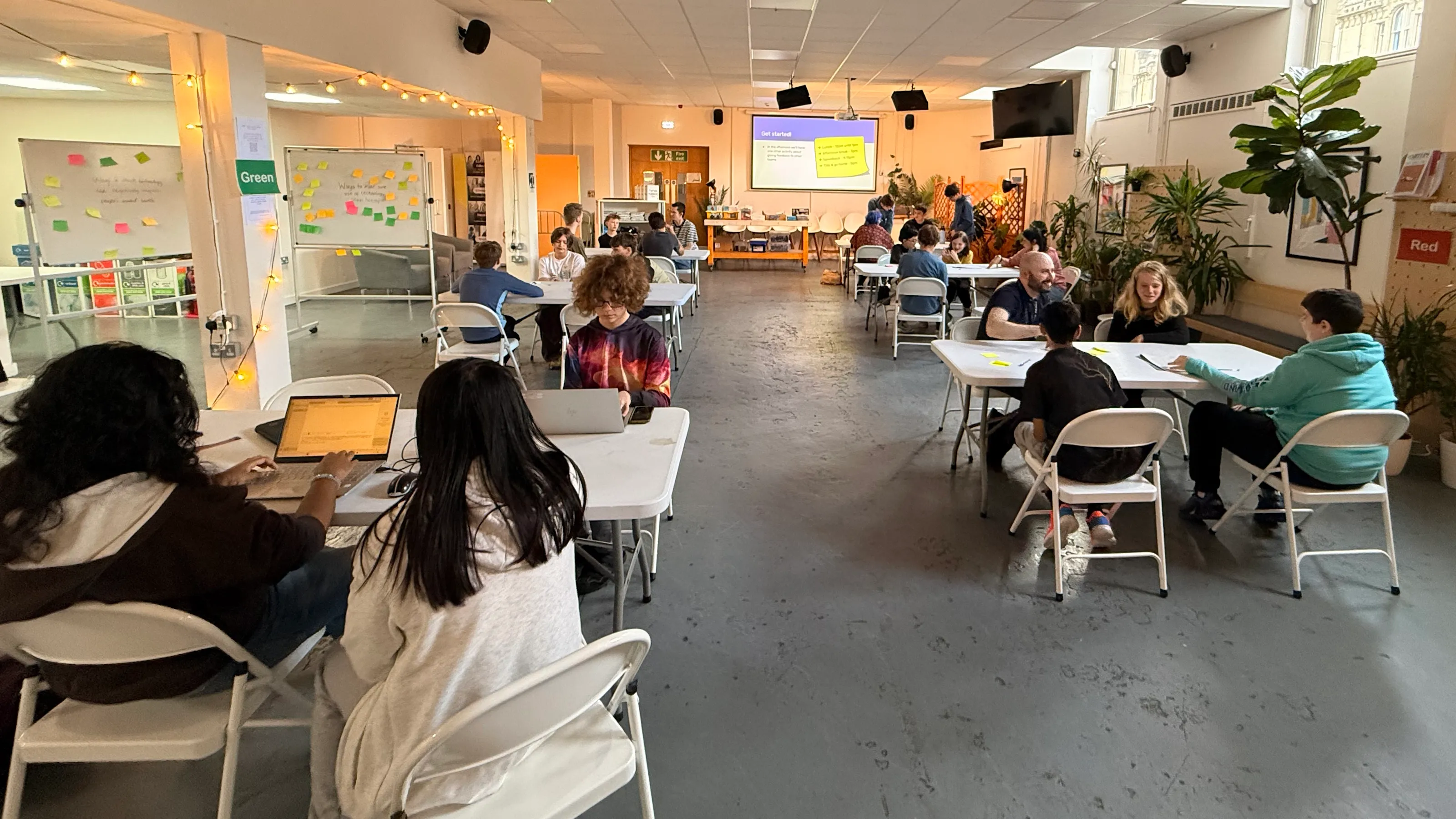 A large room filled with six teams of young people working on laptops on large tables