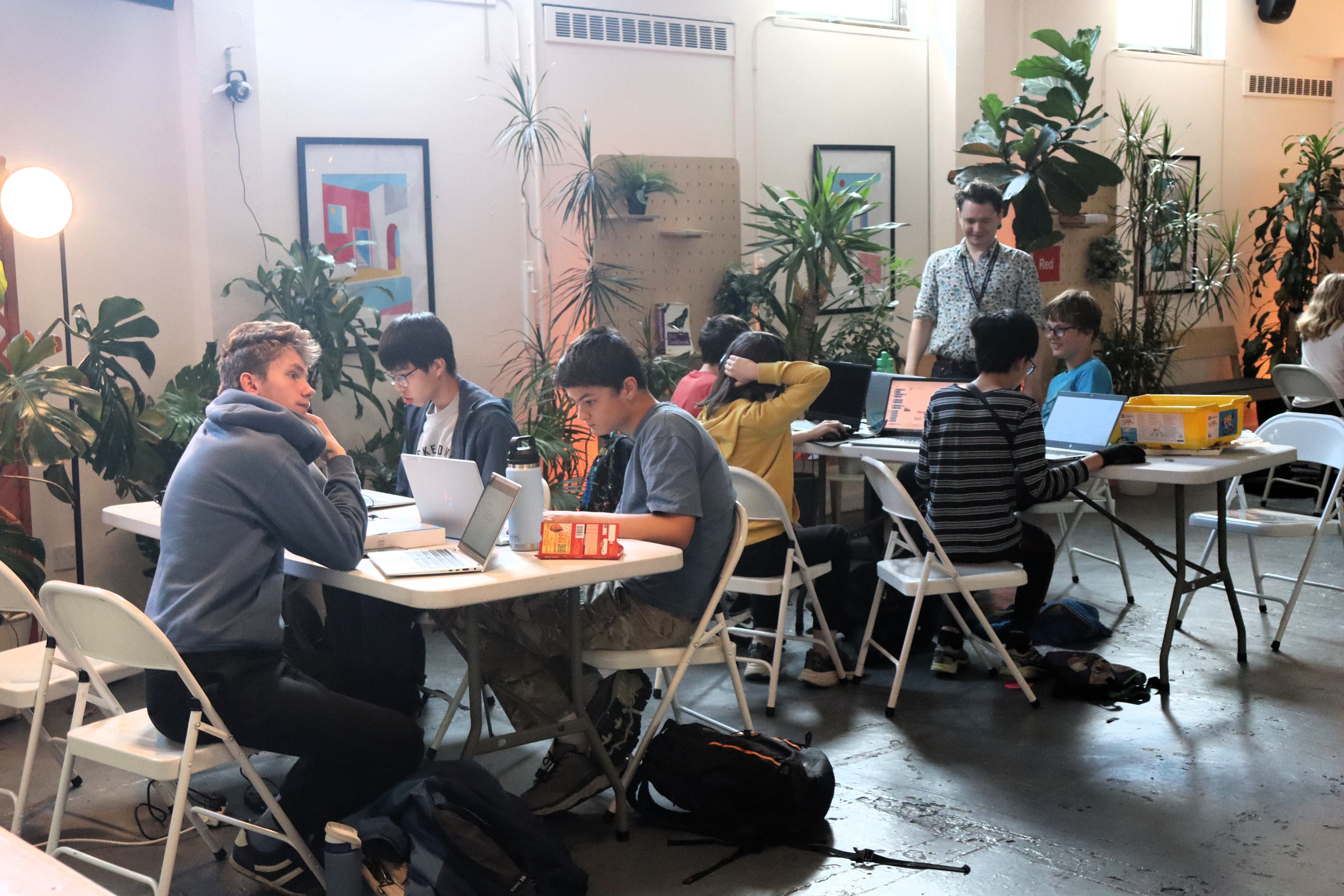 Teams of young people sitting at large tables, working on laptops